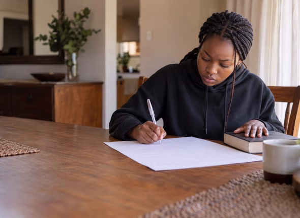 A South African Grade 12 learner writing from memory onto a blank page at the dining table, her textbook closed and face down under her other hand