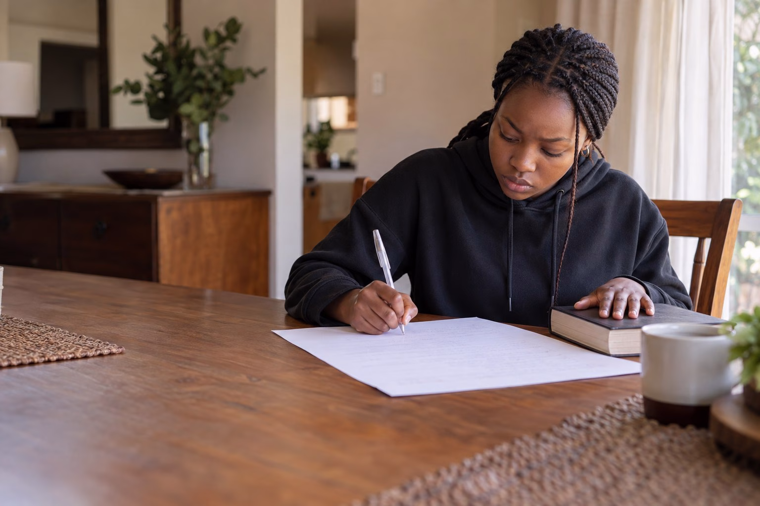 A South African Grade 12 learner writing from memory onto a blank page at the dining table, her textbook closed and face down under her other hand