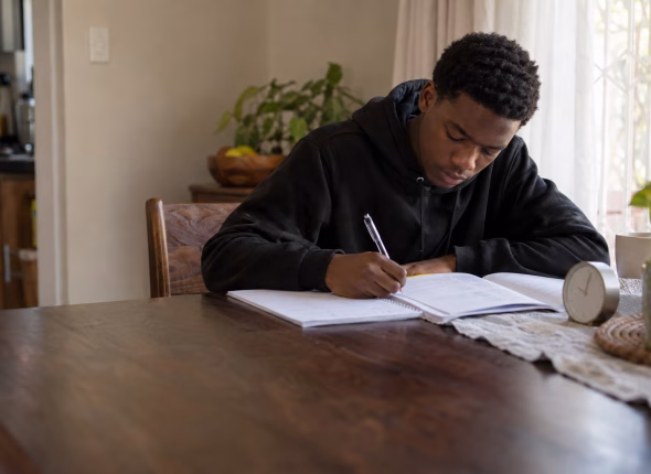 A South African Grade 12 learner writing a past paper against the clock at the dining table, a question booklet open in front of him