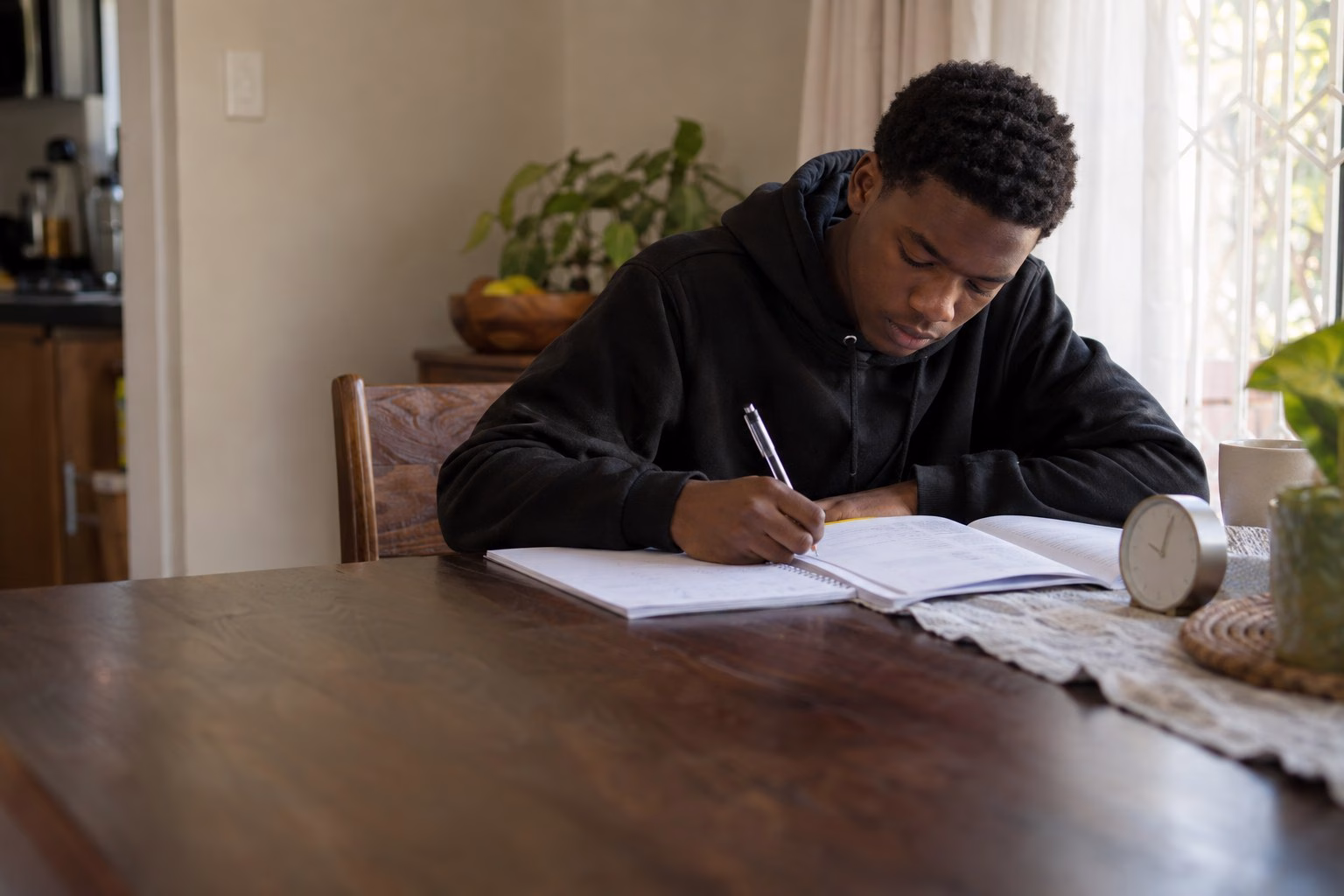 A South African Grade 12 learner writing a past paper against the clock at the dining table, a question booklet open in front of him
