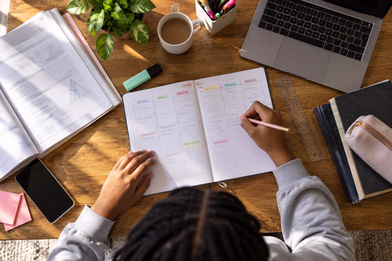 Overhead view of a learner filling in a colour-coded weekly study timetable in a notebook, with a textbook, laptop, ruler and highlighters on the desk