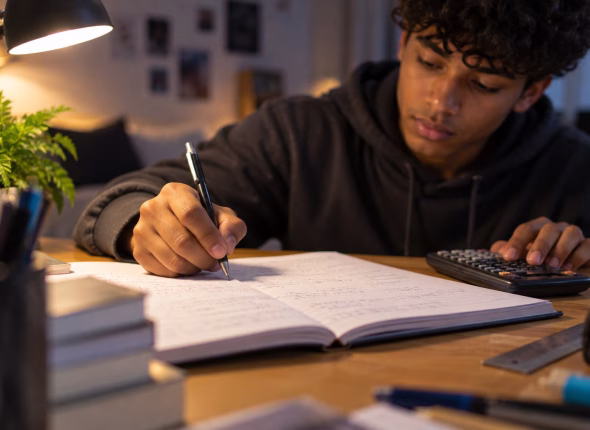 A South African Grade 12 learner working a mathematics problem by hand in an exercise book under a desk lamp, one hand on a scientific calculator