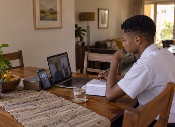 A South African learner at a dining table during an online tutoring lesson, laptop open beside a phone propped up as a mobile data backup