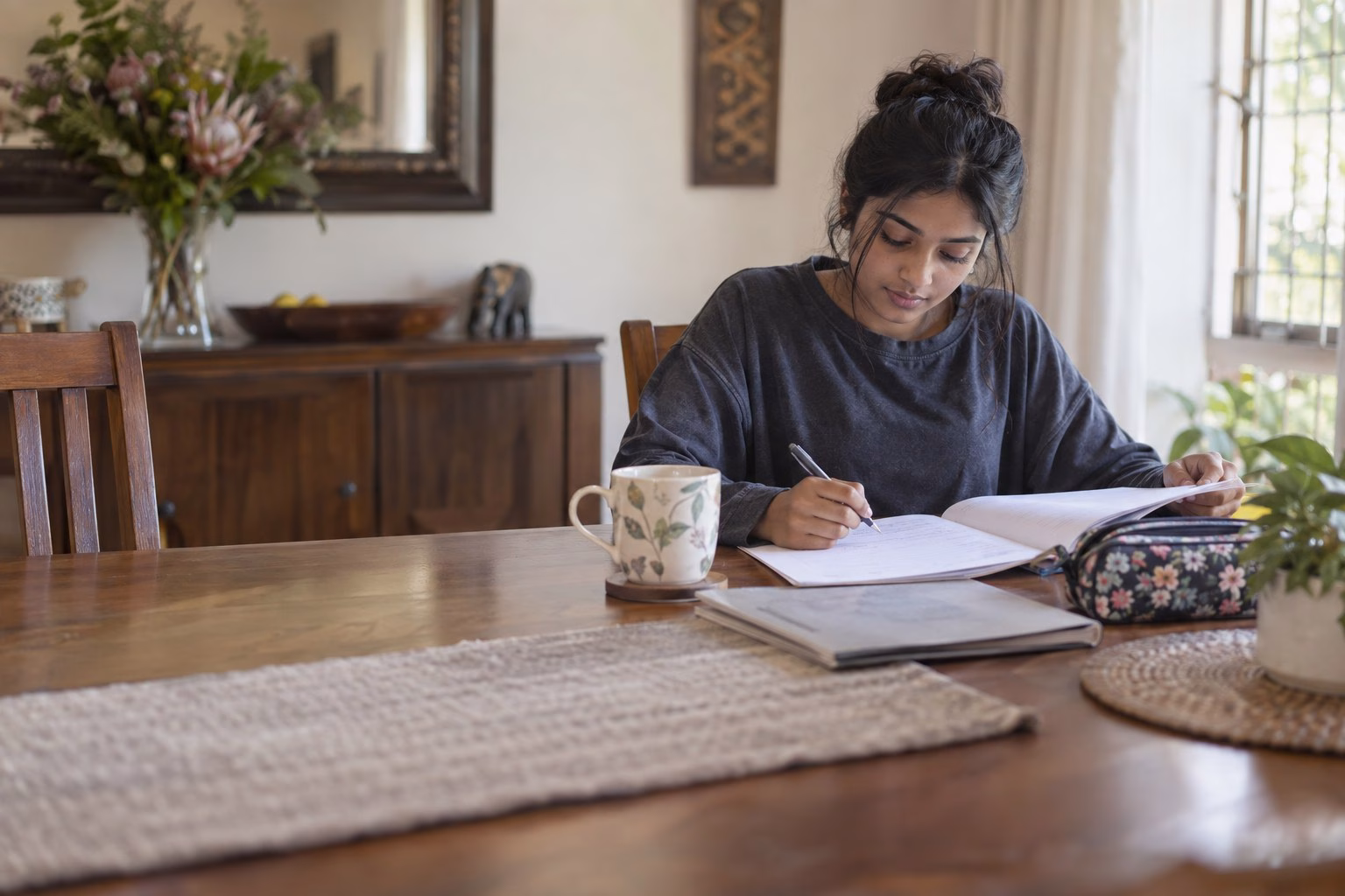 A South African Grade 12 learner working through past papers at the dining table during the September school holiday, with a mug and a wall calendar behind her