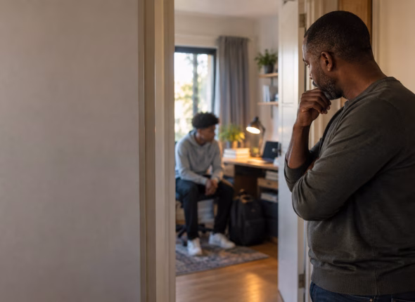 A South African father standing in a hallway looking thoughtfully towards his teenage son's bedroom, where the son sits on the bed with a closed schoolbag beside him