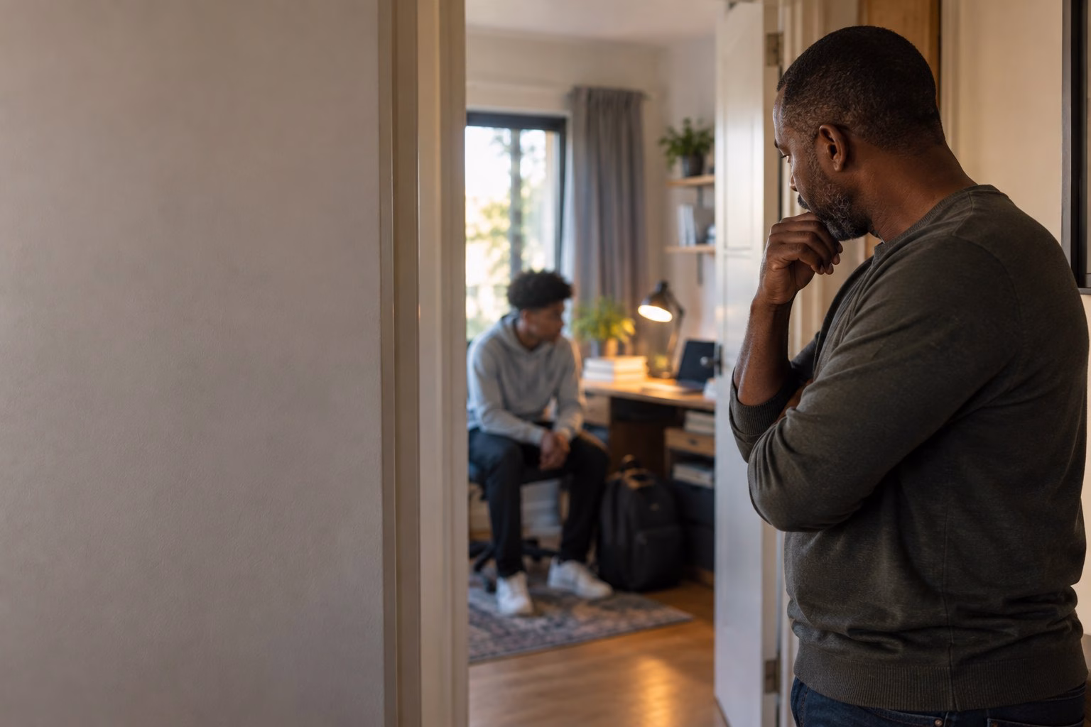 A South African father standing in a hallway looking thoughtfully towards his teenage son's bedroom, where the son sits on the bed with a closed schoolbag beside him
