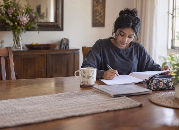 A South African learner writing by hand in an exercise book at a dining table, turning the page of a past paper, with a mug and a pencil case beside her