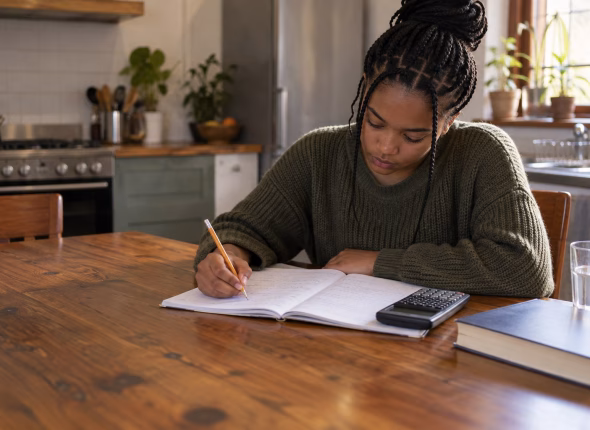 A South African school-leaver working through mathematics problems by hand in an exercise book at a kitchen table, with a scientific calculator beside the book