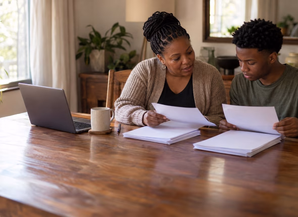 A South African mother and her son, a school-leaver, reading printed sheets together at a dining table with two neat stacks of paper and a laptop beside them